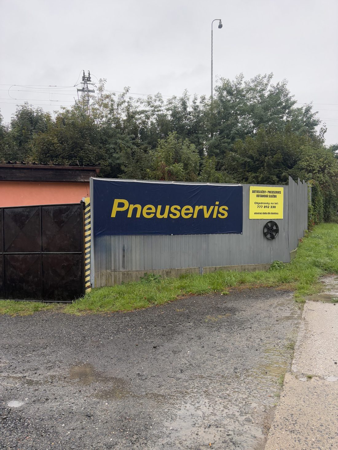 Tmavomodrá cedule s nápisem Pneuservis na plechovém plotu u vjezdu, vedle menší žlutá cedule
